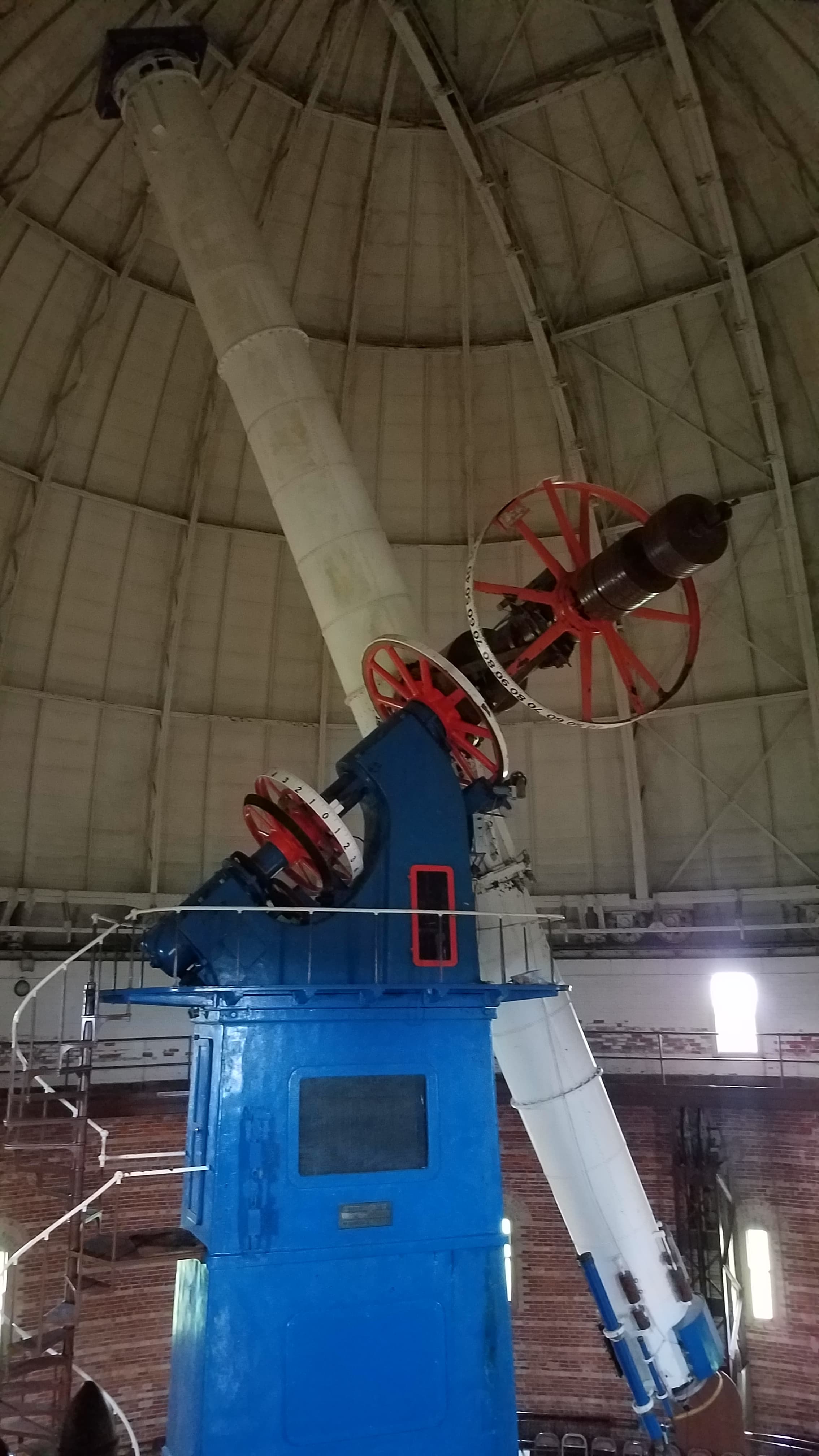 a long white telescope on a large blue pier inside a dome. There is spiral staircase on the left leading up to a platform on the pier, and there are also orange wheels and brown weights through the pier and perpendicular to the telescope.