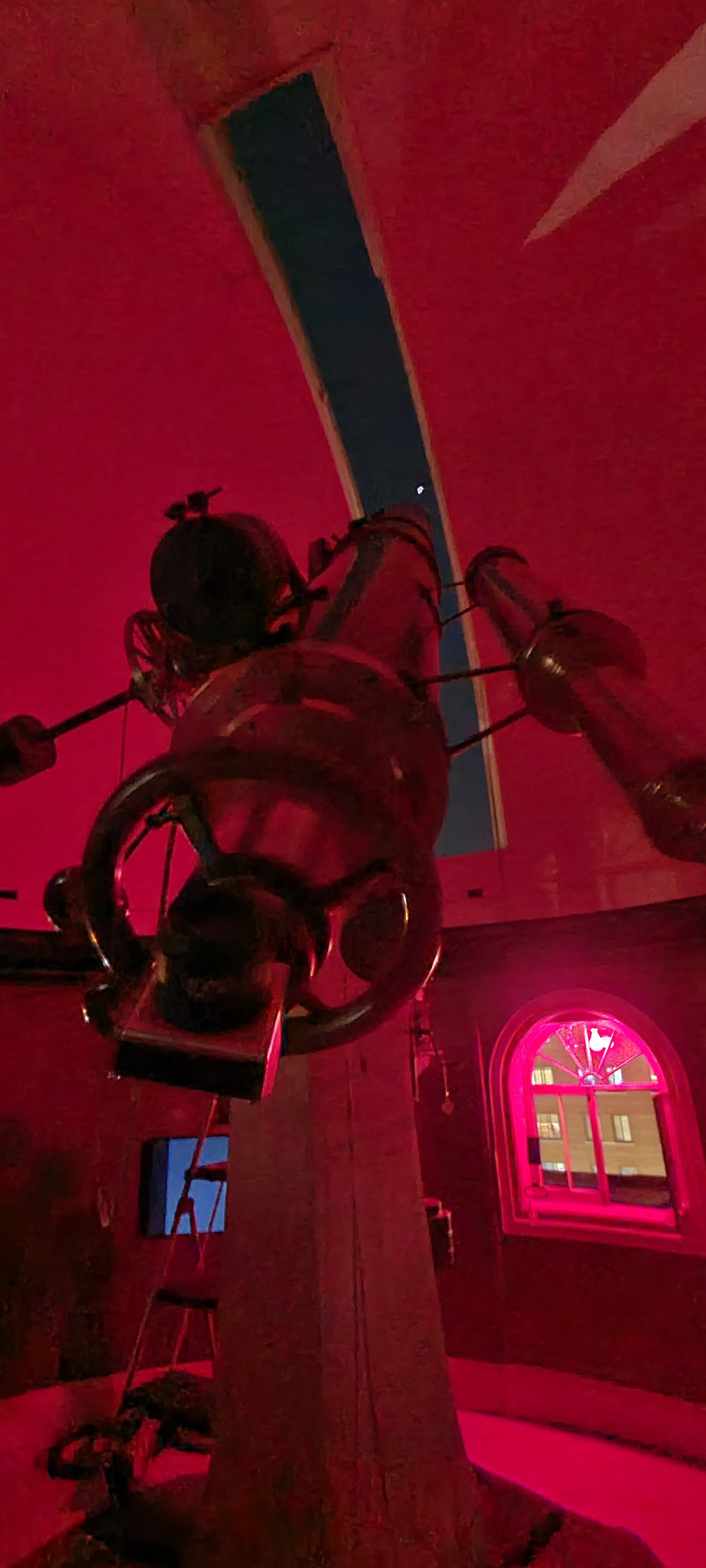 View looking down the barrel of the a telescope that is pointed at the moon that can be seen through a narrow dome slit. Everything is bathed in red light from a red lightbulb that is hanging by a window.