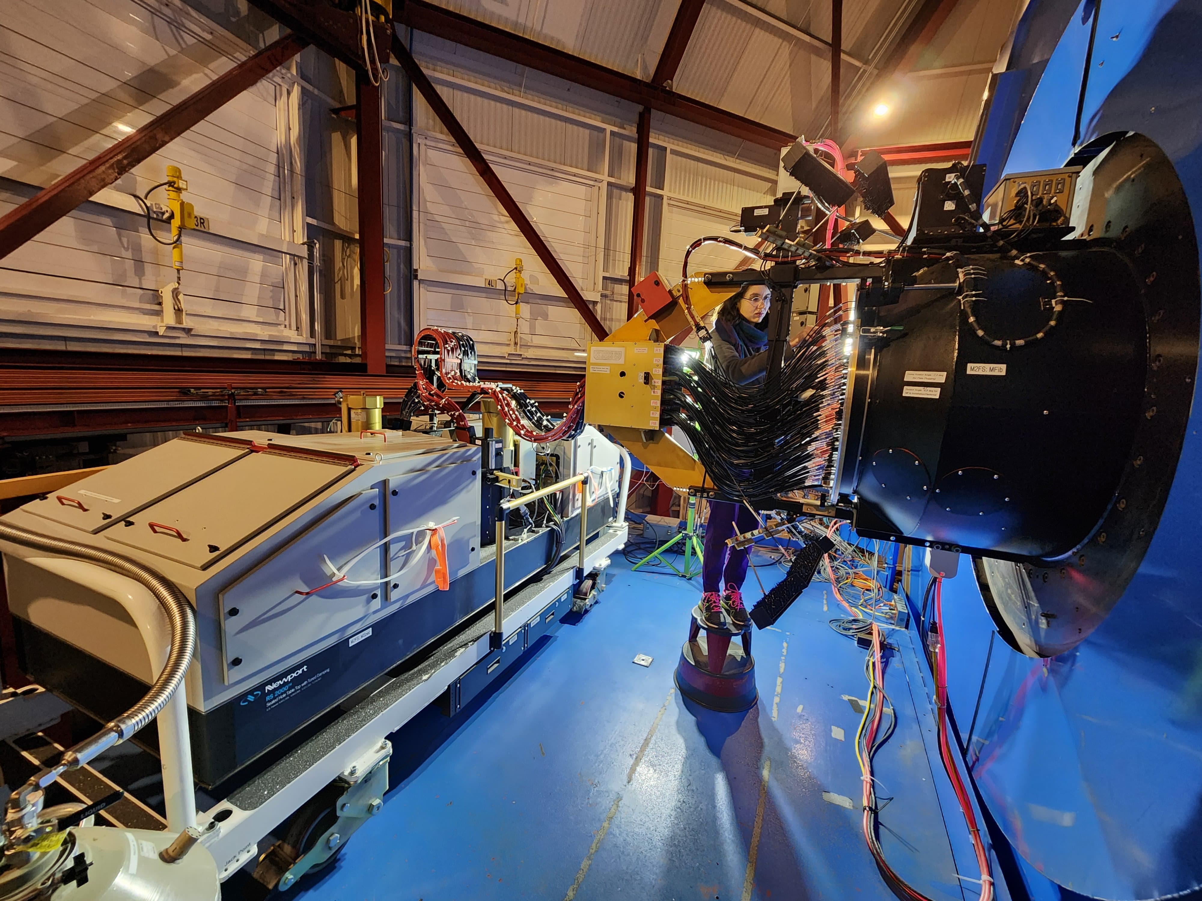 A girl with glasses and bundled in a scarf, fleece, and long purple pants stands on a stepstool behind a big bundle of fiber optic cables connected from a plate to a spectrograph. She is plugging one of the fibers in.