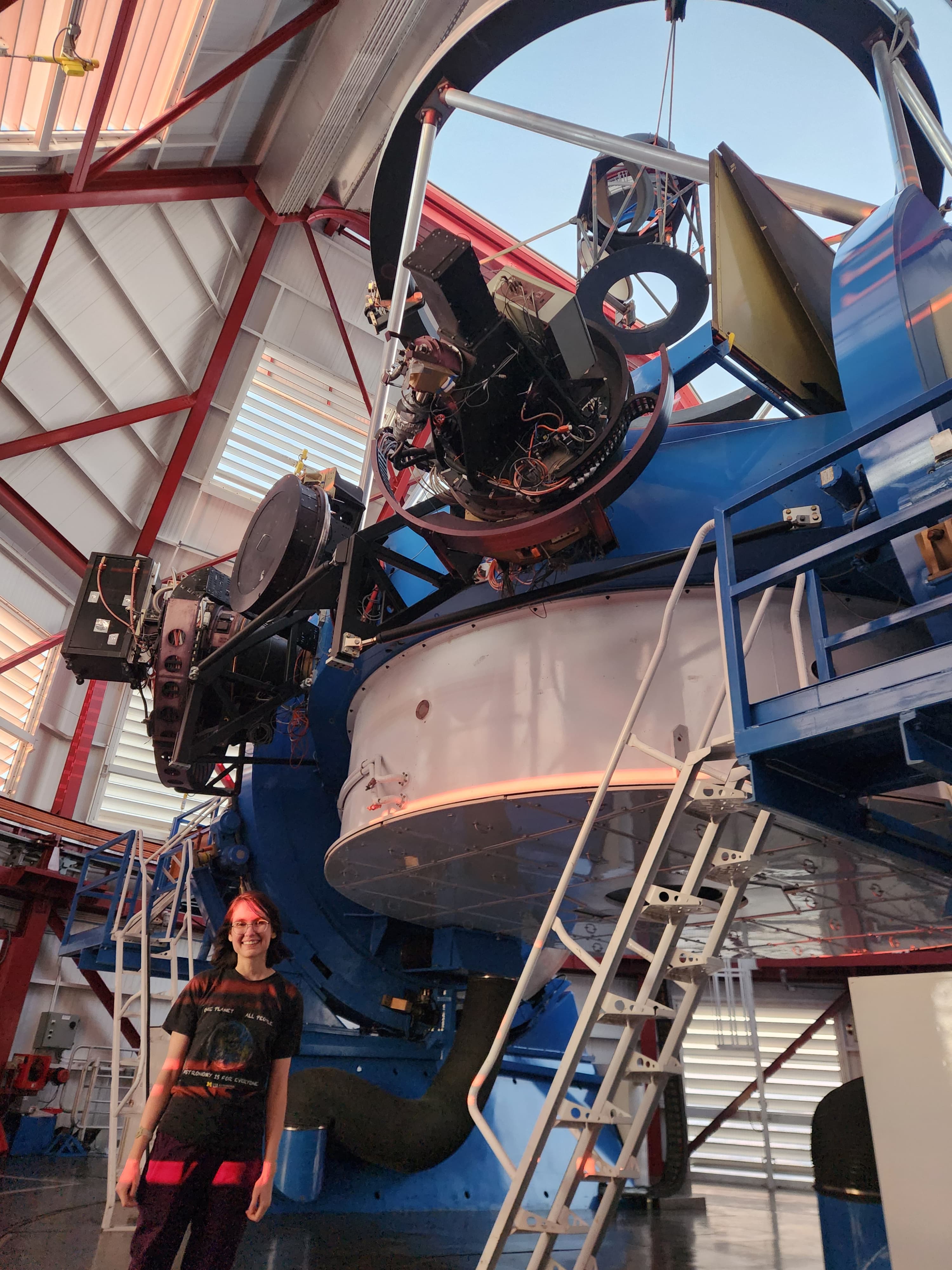A girl standing next to a giant reflecting telescope inside a dome that is open to a light blue sky.