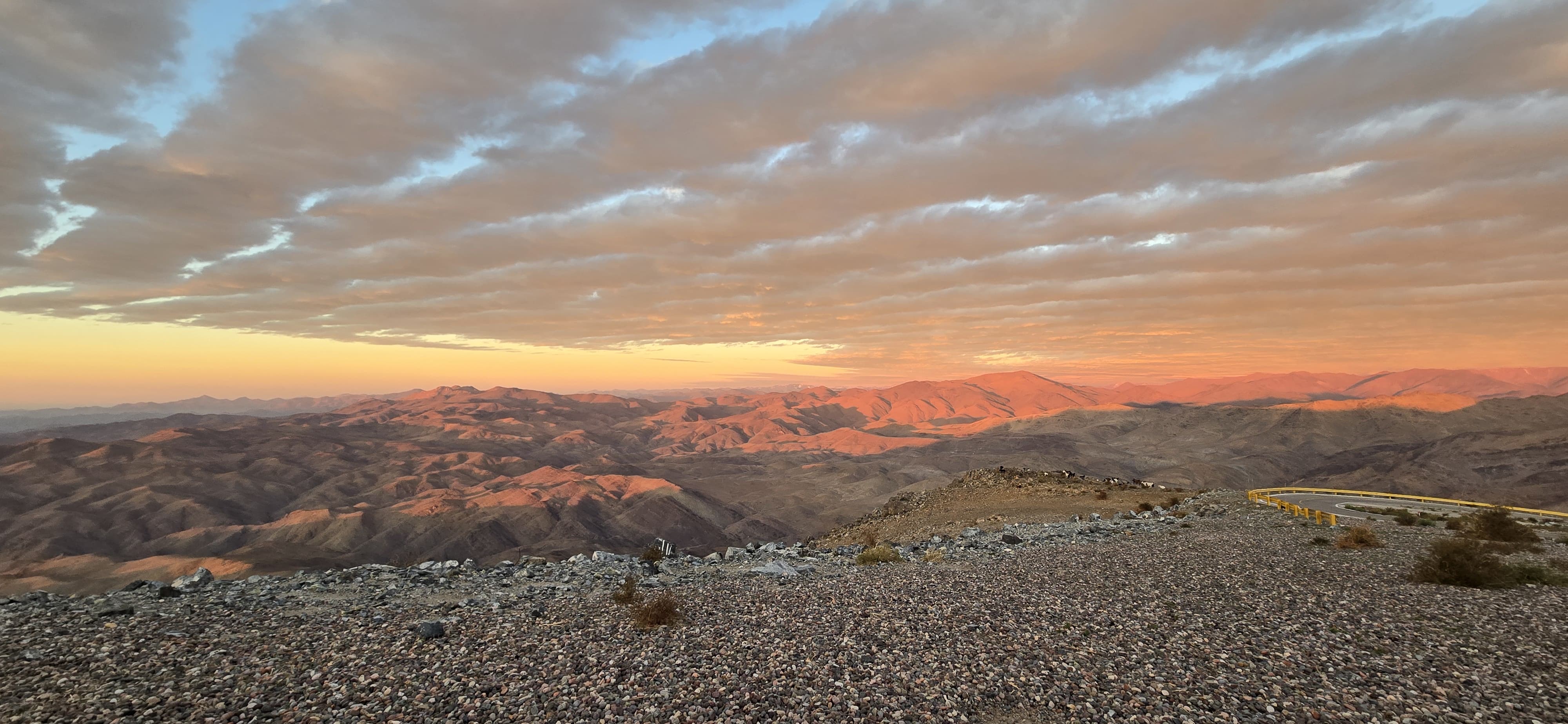 Sunset-colored mountains stretch out below a cloudy sky. The foreground is rocky land with a road curving on the right side. In the distance on an outcropping is a herd of goats.