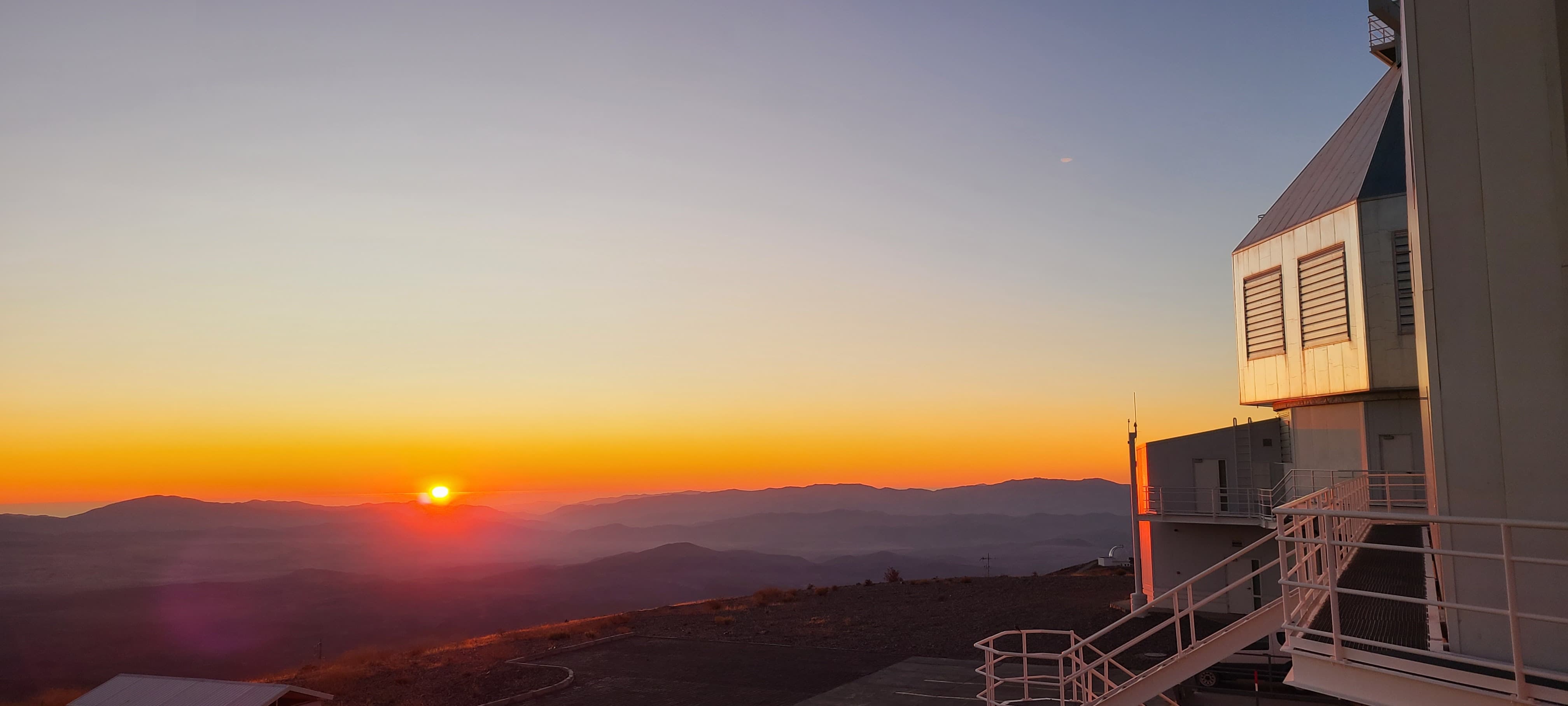 A sun setting behind a mountain range with a metal telescope dome to the right.